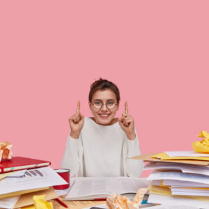 Positive European woman with glad facial expression, dressed in white casual clothes, shows direction upside, uses paper documents, has toothy smile, isolated over pink background, reads books