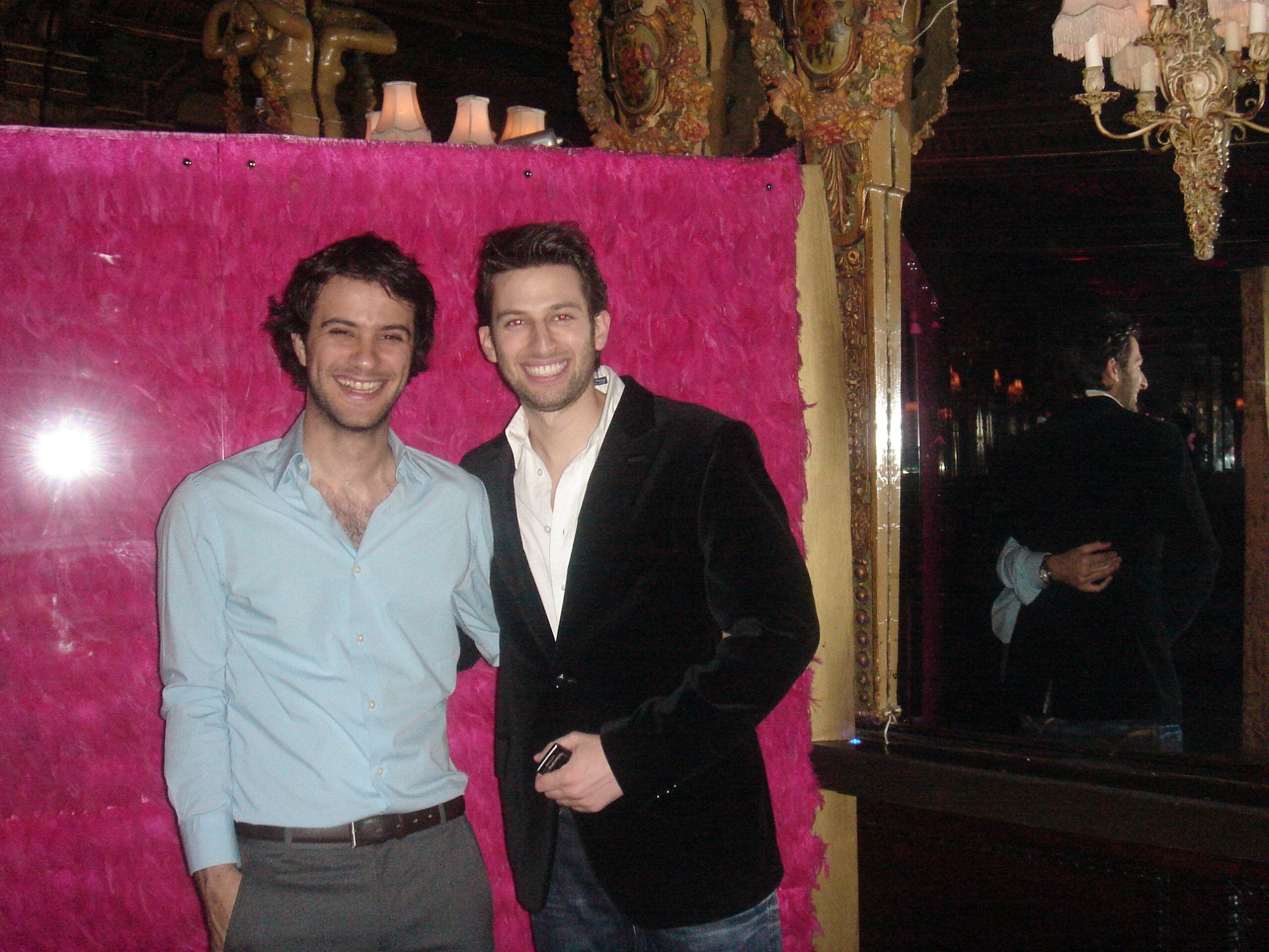 Two friends smiling in front of a pink feather wall at the Café Royal